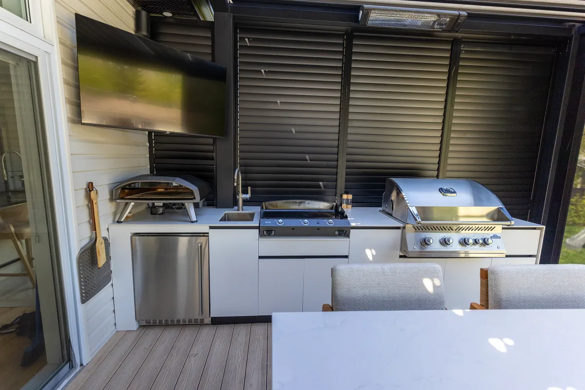 White outdoor kitchen with pizza oven, plancha, sink and built-in barbecue, set against a louvred privacy wall