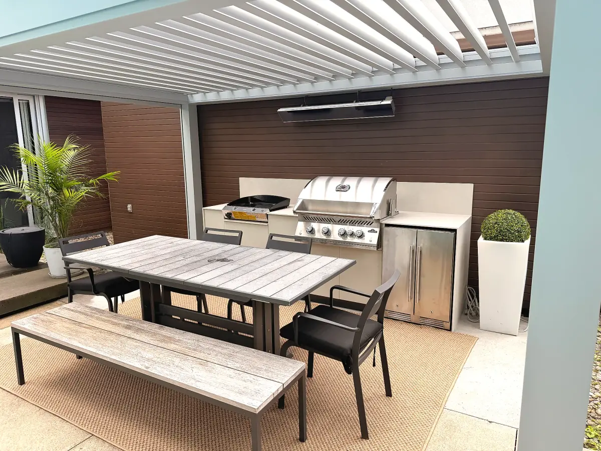 Outdoor kitchen with cream cabinets, built-in plancha and barbecue, under a bioclimatic pergola, with a dining table