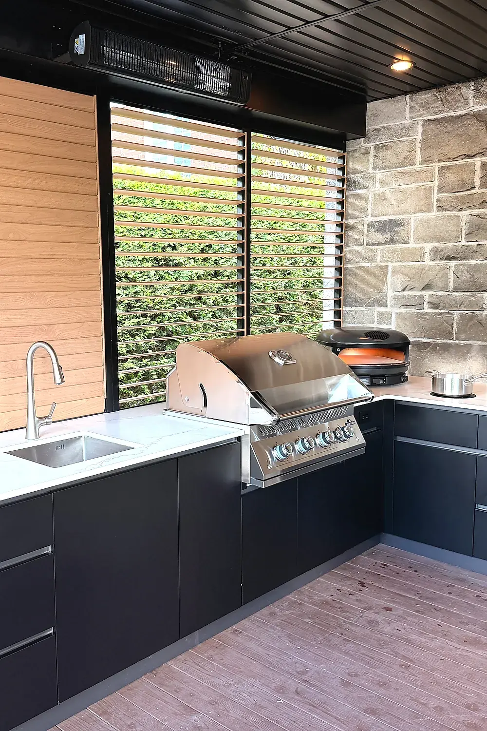 L-shaped outdoor kitchen with black cabinets and white sintered stone counters: sink, built-in barbecue and pizza oven, against a stone wall and a louvred privacy wall