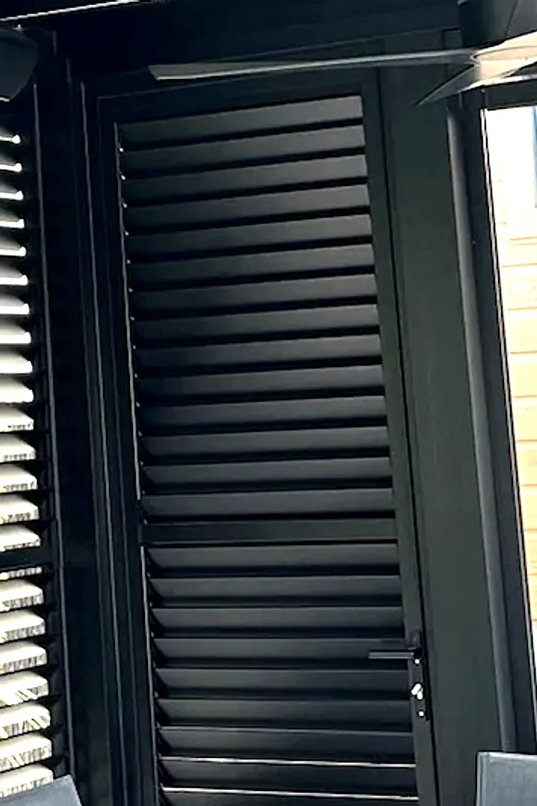 Porte en persiennes d'aluminium noir intégrée à un mur d'intimité sous une pergola : deux registres de lames séparés par une traverse, poignée à levier et cylindre de serrure sur le montant, panneau voisin ouvert à gauche