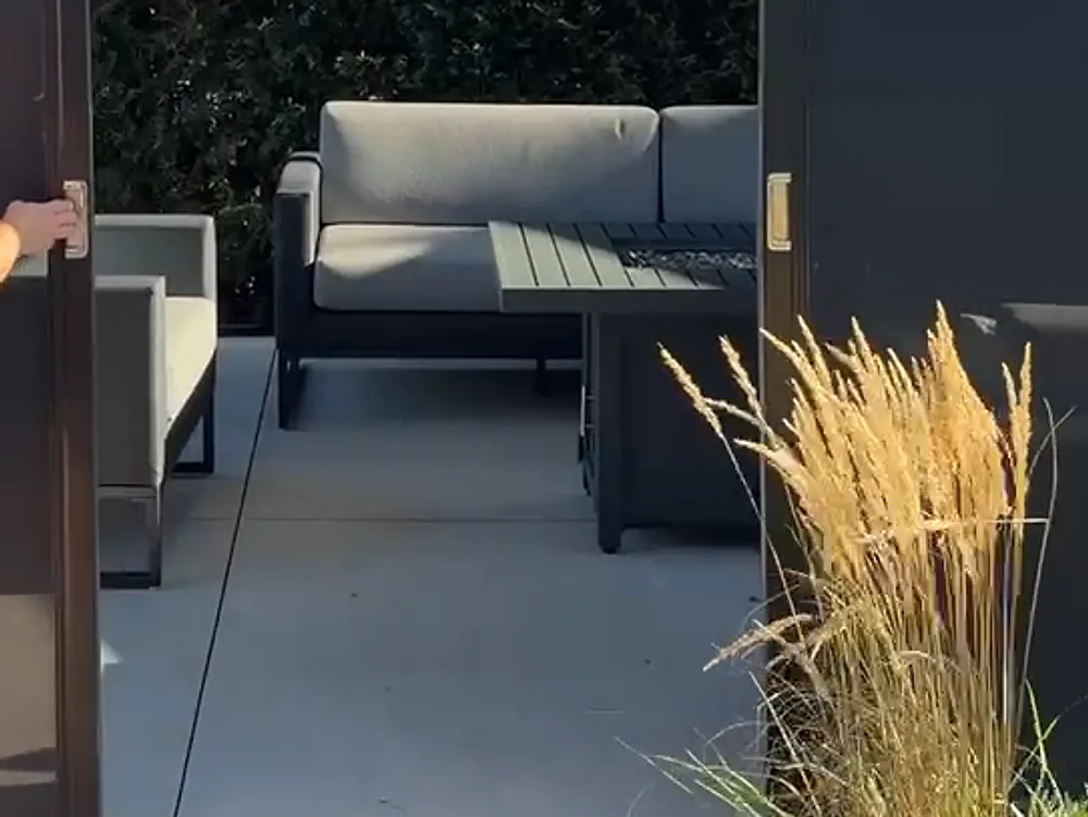 Panel of a horizontal solar shade at the end of its run, a hand on the brass handle of the operating bar, the opening clear onto the outdoor lounge and the concrete slab