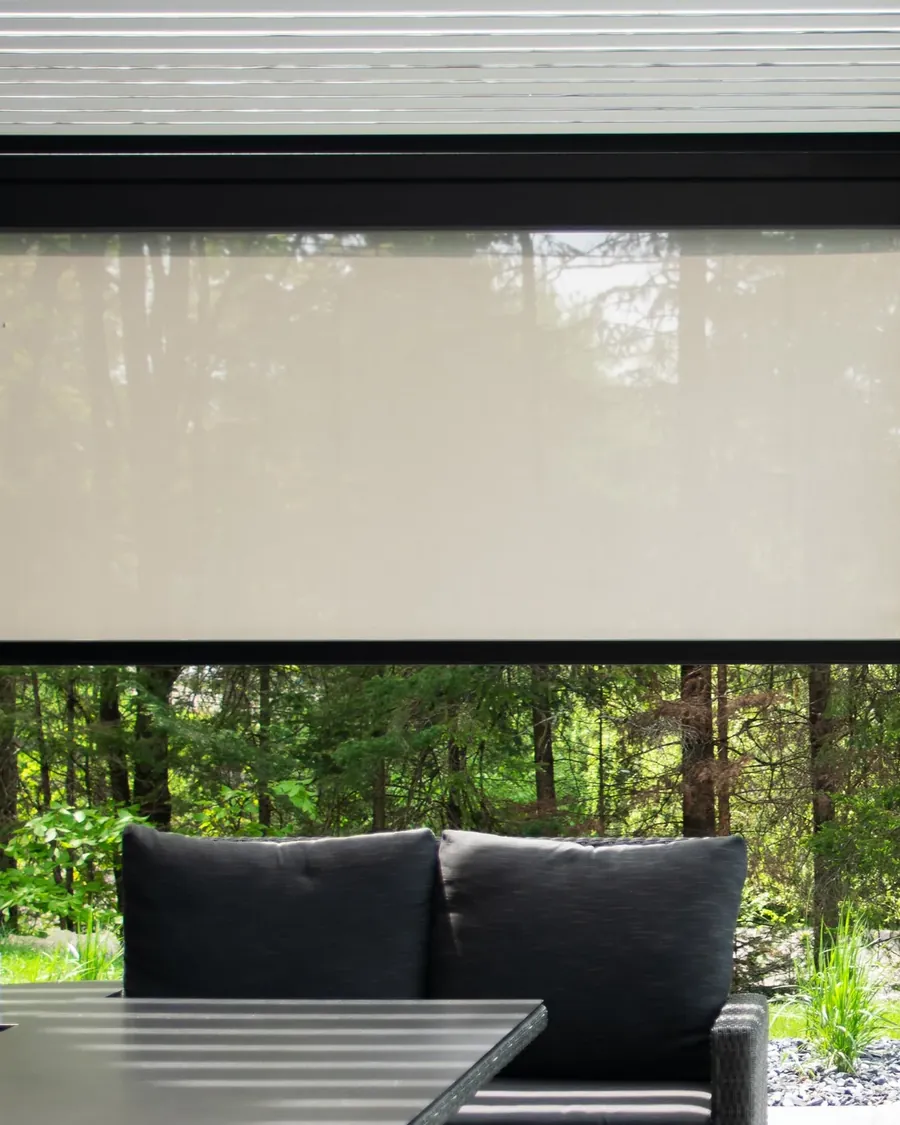Cassette d’une toile solaire posée en tête d’ouverture sous les lames d’une pergola, la toile arrêtée à mi-hauteur, salon extérieur et sous-bois en contrebas