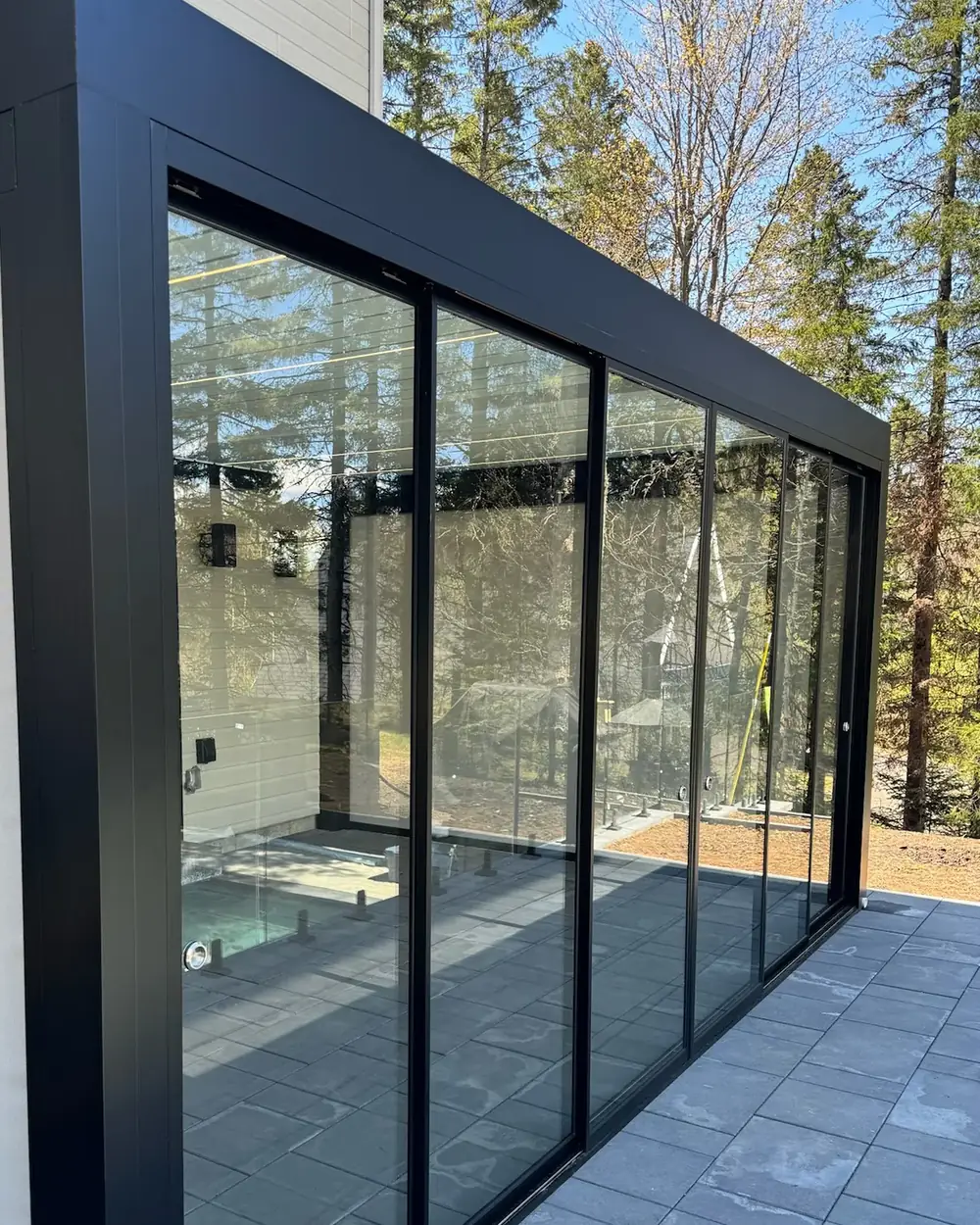 Verre coulissant à cadre noir le long d'une pergola adossée à une résidence, vu de trois quarts; le rail est visible au pied des panneaux