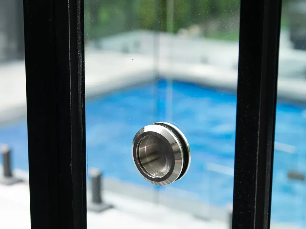 Poignée encastrée en acier inoxydable sur un panneau coulissant, entre deux montants de cadre noirs; piscine creusée à l'arrière-plan
