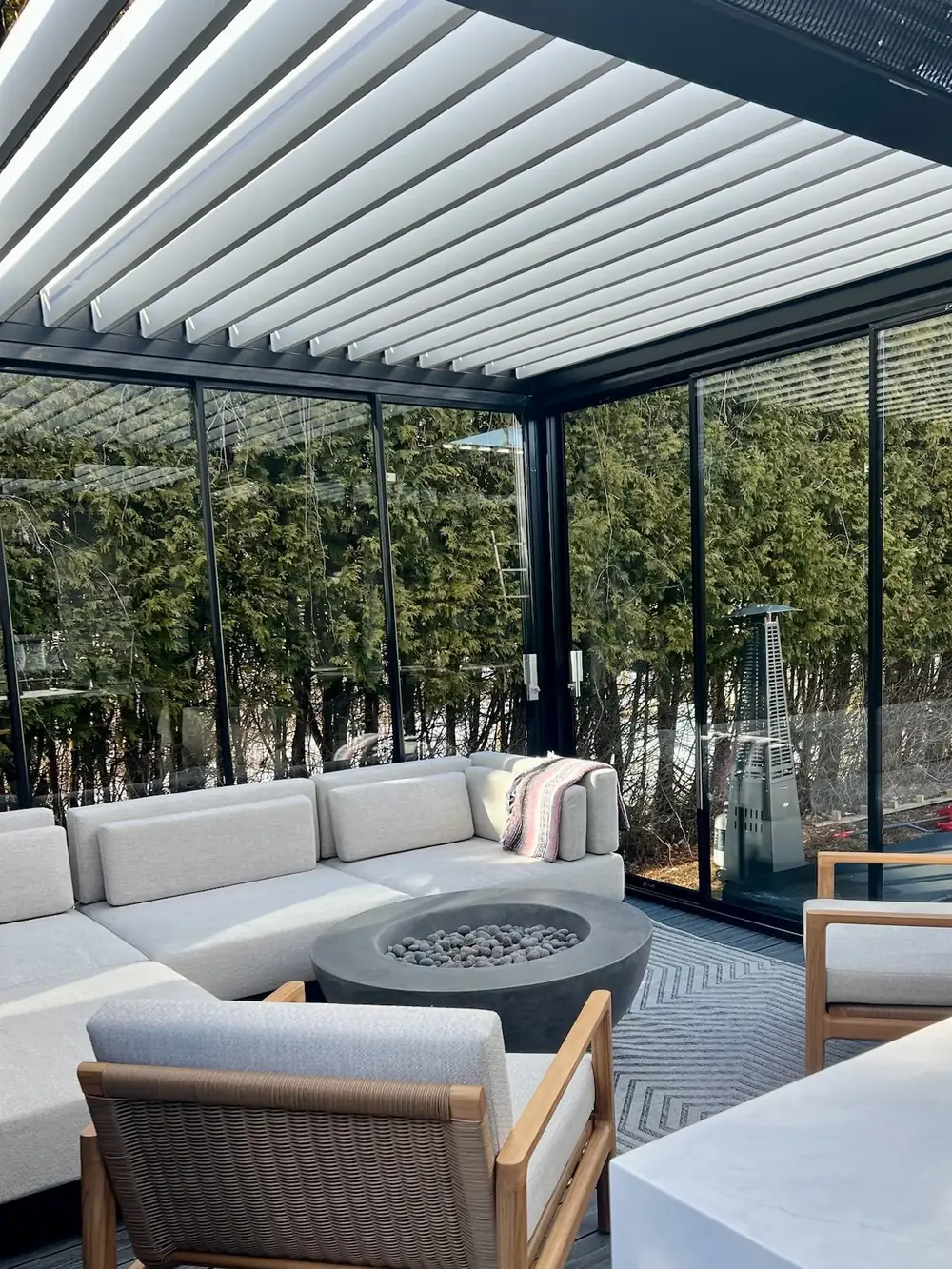 Salon extérieur sous une pergola à lames orientables blanches, fermé sur deux côtés par du verre coulissant; sofa modulaire, foyer au gaz et haie de cèdres derrière le verre