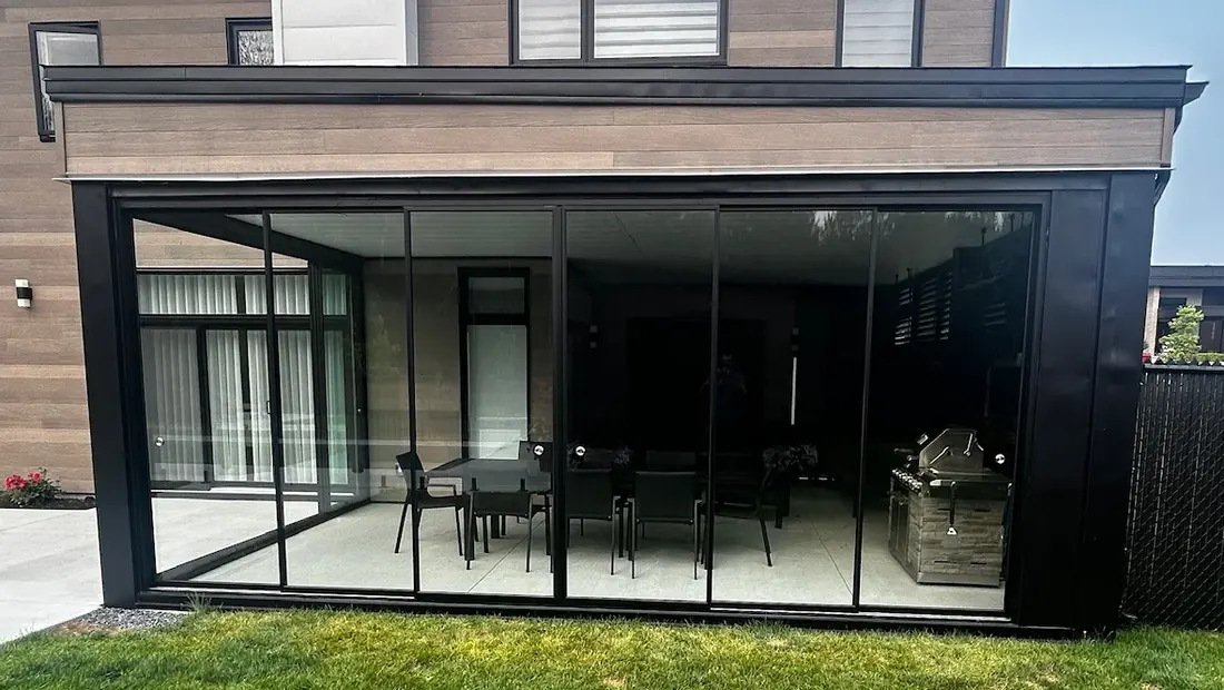 Espace couvert entièrement fermé par du verre coulissant, vu du jardin : table à manger, barbecue et mur d'intimité à lames à l'intérieur