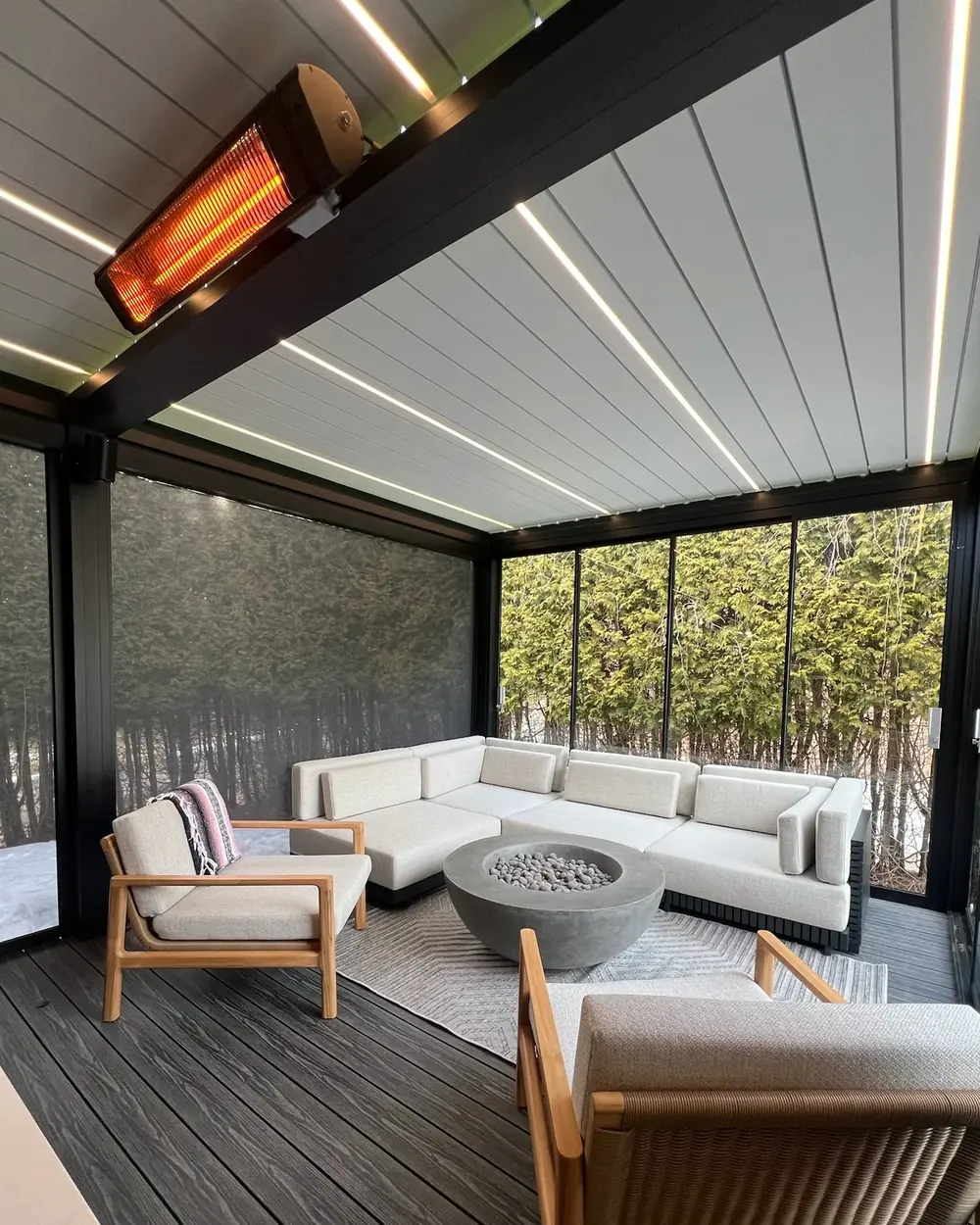 Intérieur d'une pergola fermée en fin de saison : lames blanches et éclairage LED au plafond, chauffe-terrasse allumé, toile solaire descendue à gauche, verre coulissant à droite et neige au sol dehors