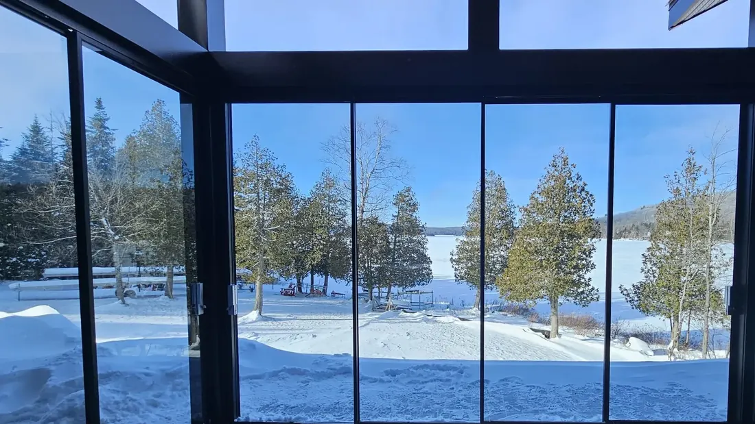 Vue sur un lac gelé et sa rive boisée à travers une paroi de verre coulissant à cadre noir