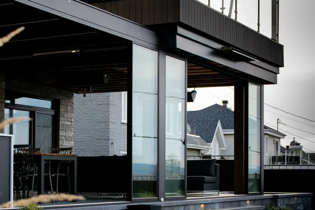 Verre coulissant fermant une terrasse sous un balcon, à la tombée du jour; comptoir-bar à l’intérieur et marches éclairées au premier plan