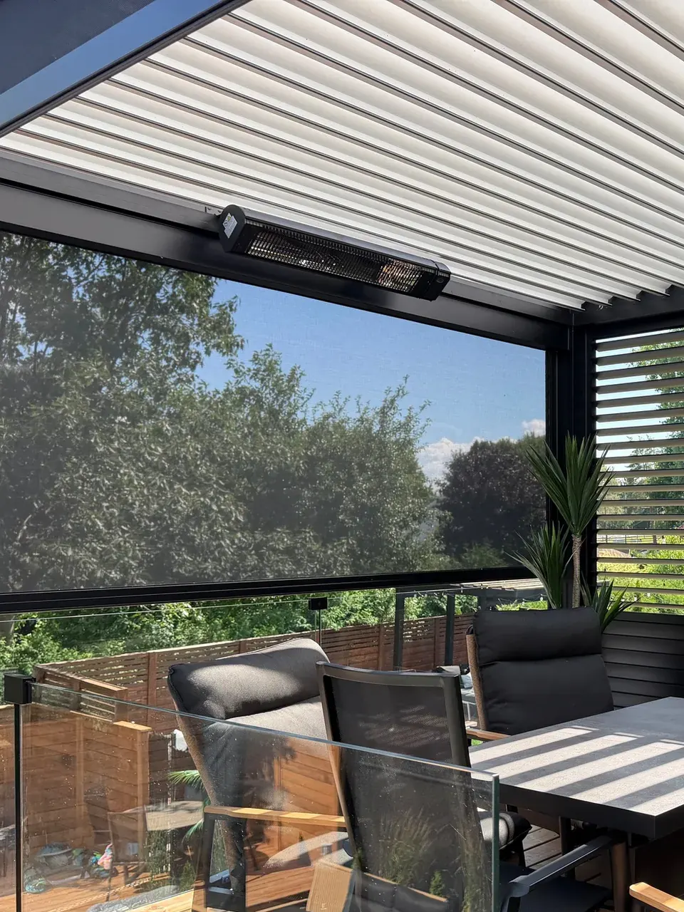 Chauffe-terrasse électrique noir fixé sous la poutre d’une pergola bioclimatique, lames blanches au-dessus, toile solaire descendue et table de terrasse en dessous