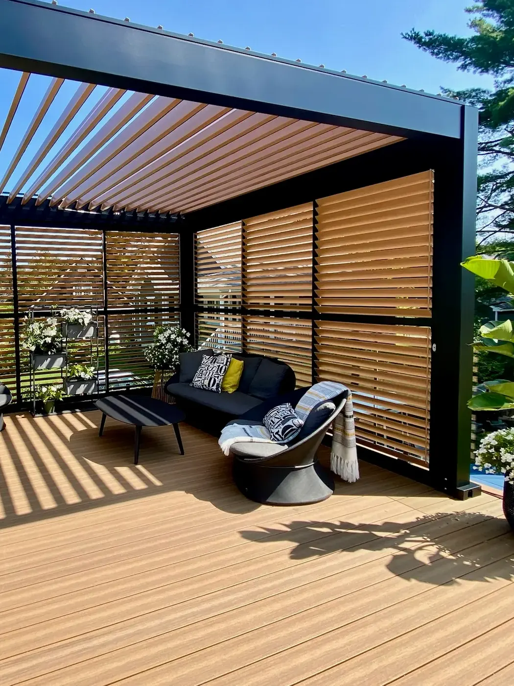 Murs d’intimité à lames au fini imitation bois fermant le fond et le côté d’une pergola en plein soleil, sofa noir et fauteuil pivotant sur une terrasse de composite