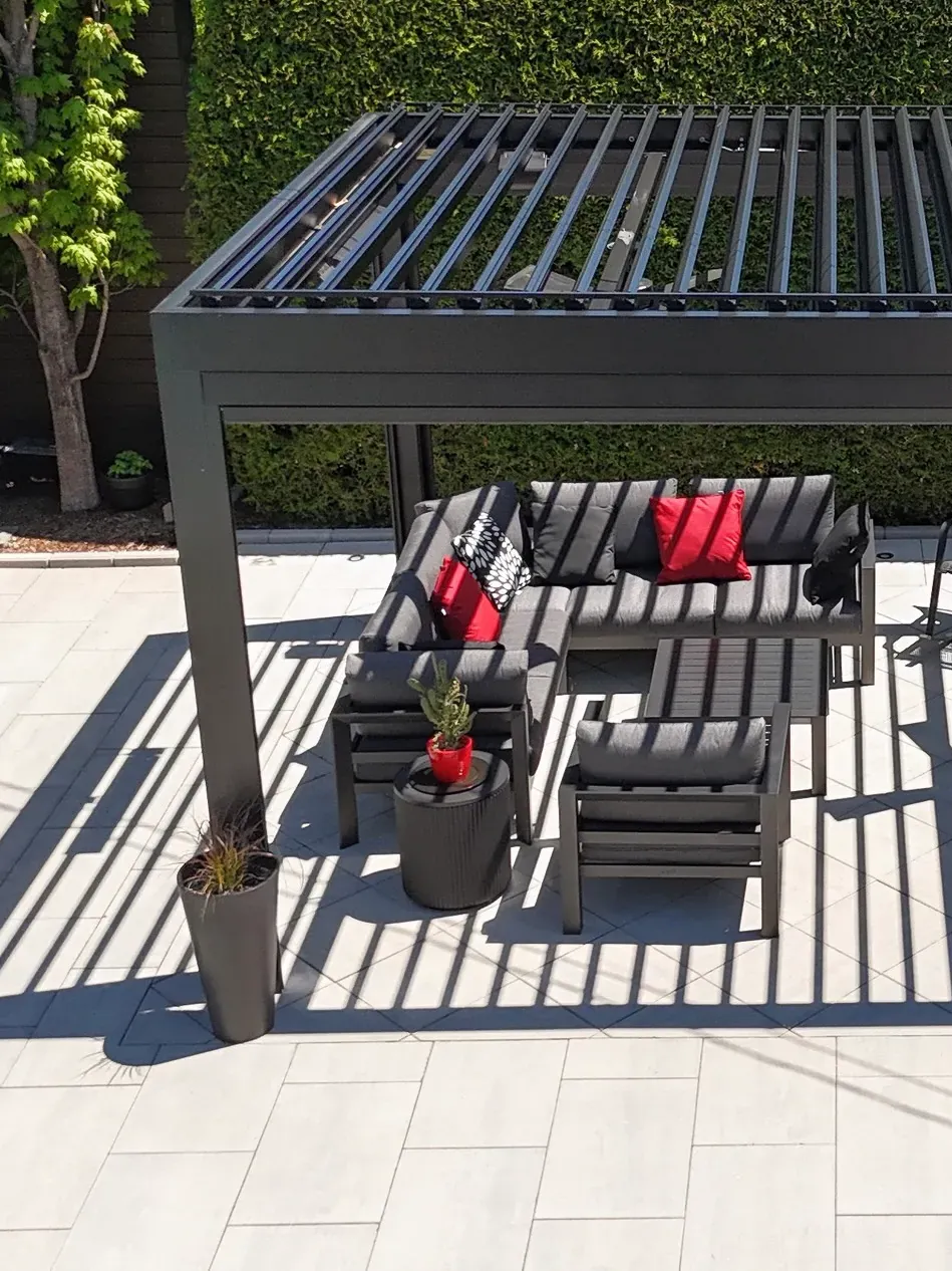 Pergola bioclimatique autoportante en aluminium noir sur une terrasse de dalles claires, lames mi-ouvertes projetant des ombres rayées, salon et salle à manger extérieurs dessous