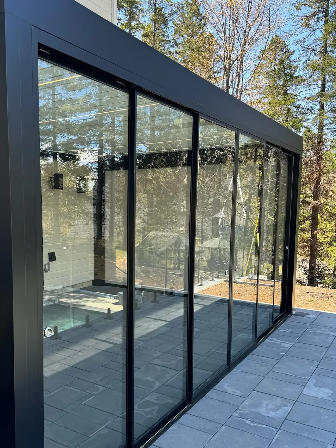 Verre coulissant à cadre noir fermant le côté d’une pergola adossée à une résidence, vu de trois quarts; dalles de pierre au sol et boisé au travers du verre