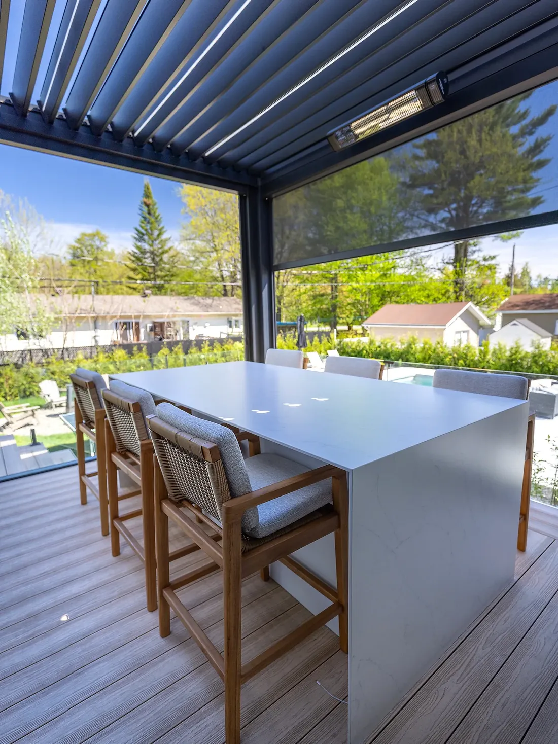Îlot-bar de cuisine extérieure en pierre frittée blanche avec tabourets en teck, sous une pergola bioclimatique à lames orientables équipée d’un chauffe-terrasse