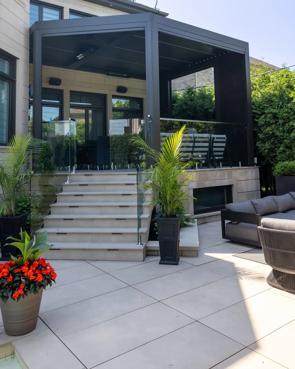 Pergola bioclimatique en aluminium noir adossée à une résidence de pierre claire : colonnes, poutre, verre coulissant et murs d’intimité au-dessus d’un escalier et d’une terrasse dallée