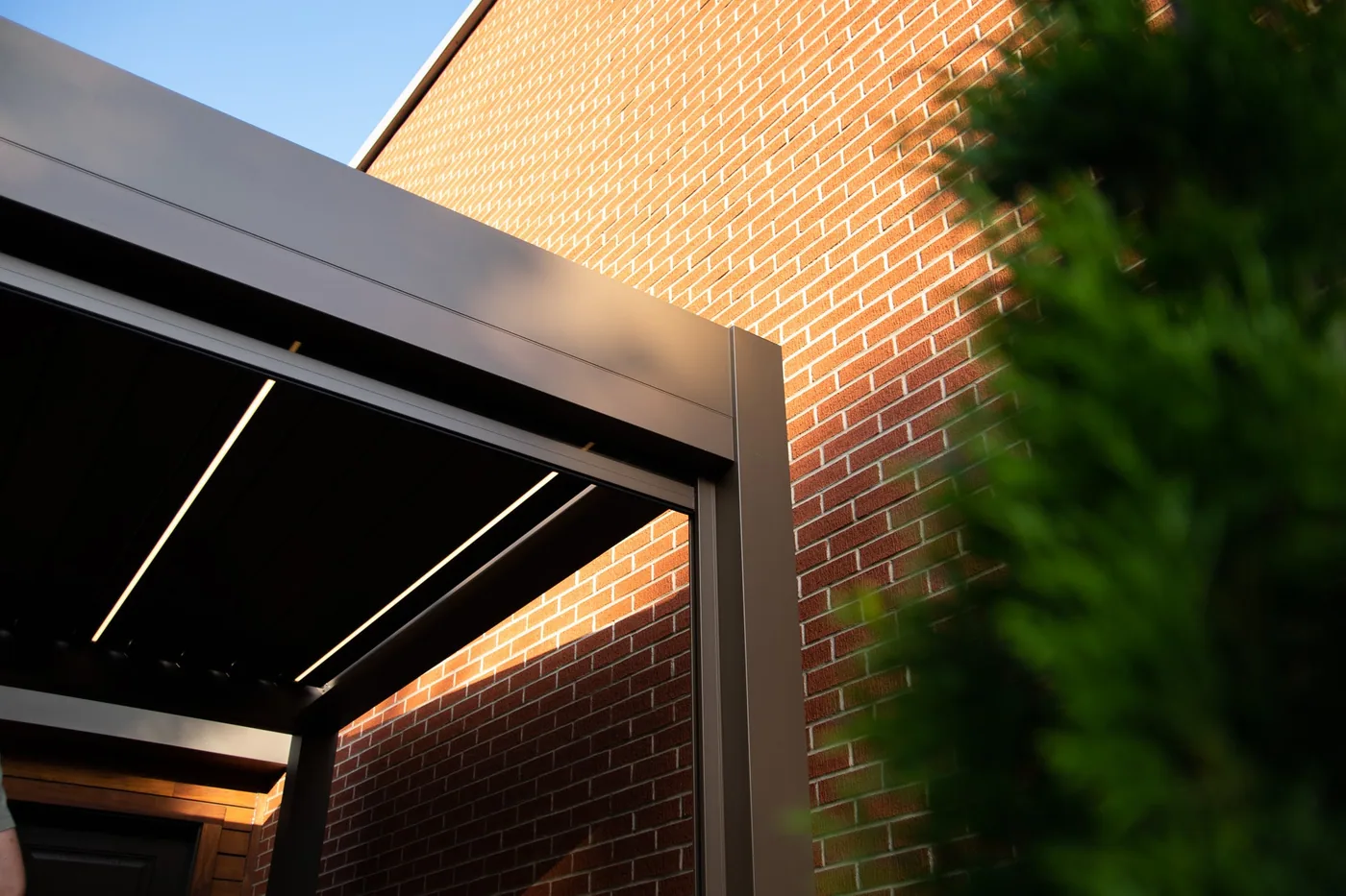 Gros plan sur la structure en aluminium d’une pergola adossée à une résidence de brique, éclairage LED intégré aux lames.