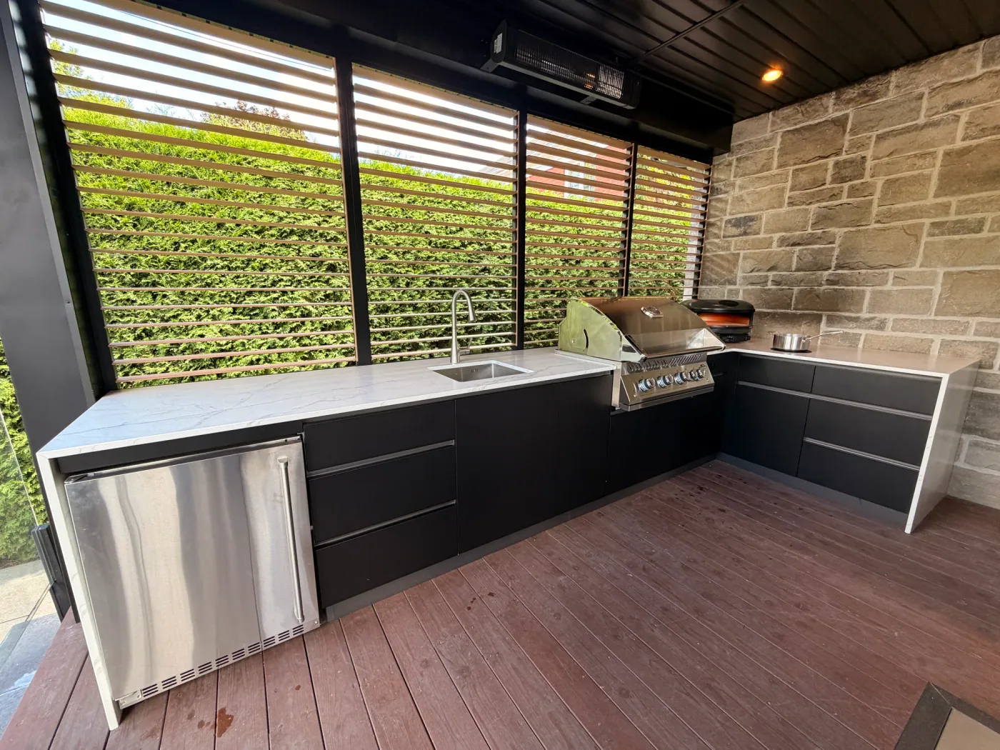 La longue ligne de la cuisine : réfrigérateur sous comptoir, évier et barbecue encastré, devant le mur à lamelles ouvert