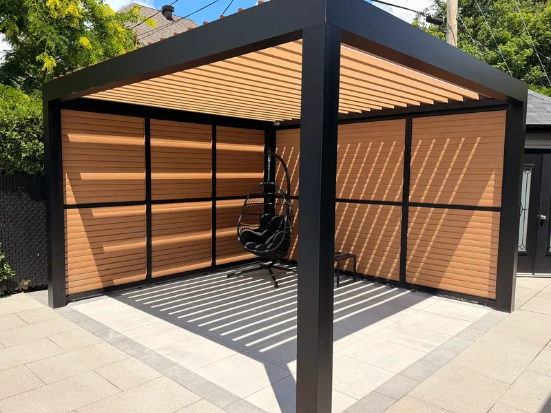 Freestanding bioclimatic pergola with wood-finish adjustable louvres, closed off by privacy walls in the same profile, on a paved terrace