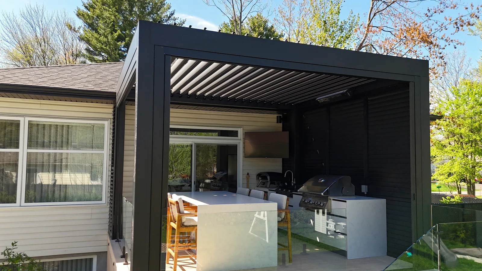 Pergola bioclimatique noire adossée à la maison à Terrebonne, à la hauteur du bord de toit, avec cuisine extérieure et mur d’intimité en persiennes
