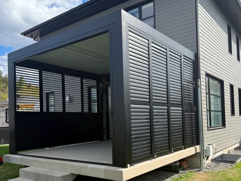 Pergola bioclimatique adossée à une maison, fermée sur le côté par un mur d’intimité en persiennes d’aluminium noir, sur une terrasse de béton