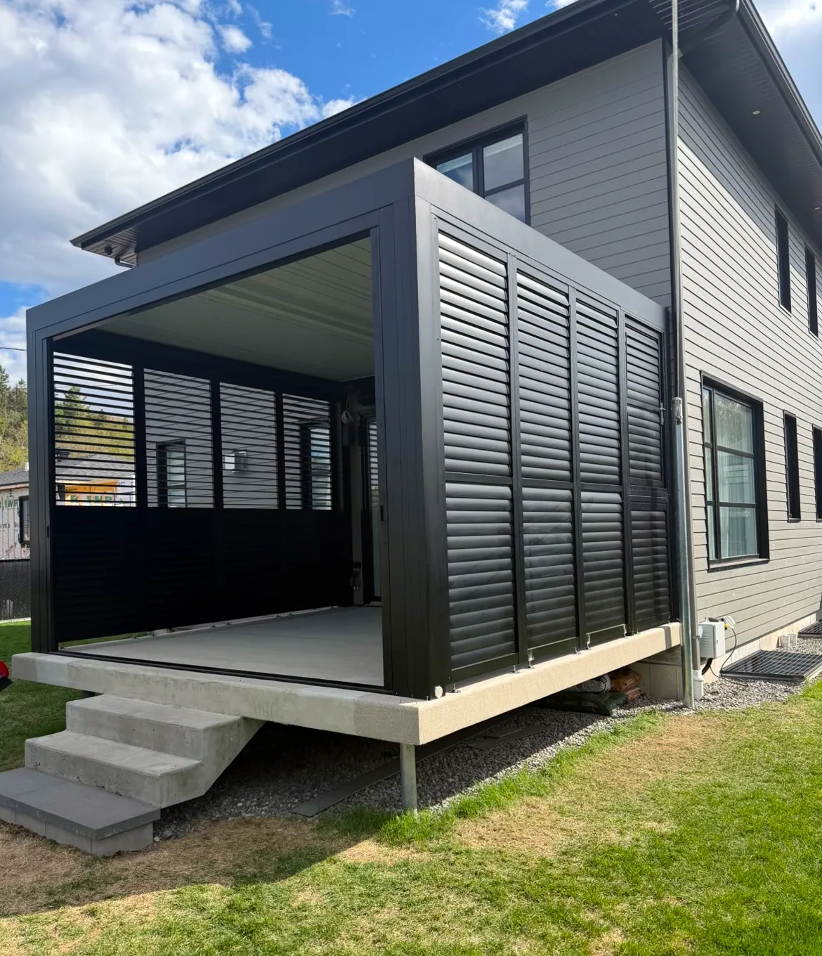Bioclimatic pergola against a house, closed on one side by a black aluminum louvred privacy wall, on a concrete terrace