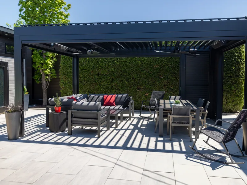 Pergola bioclimatique Capri noire au-dessus d’une terrasse de dalles, entre la maison et une haie de cèdres, à Laval