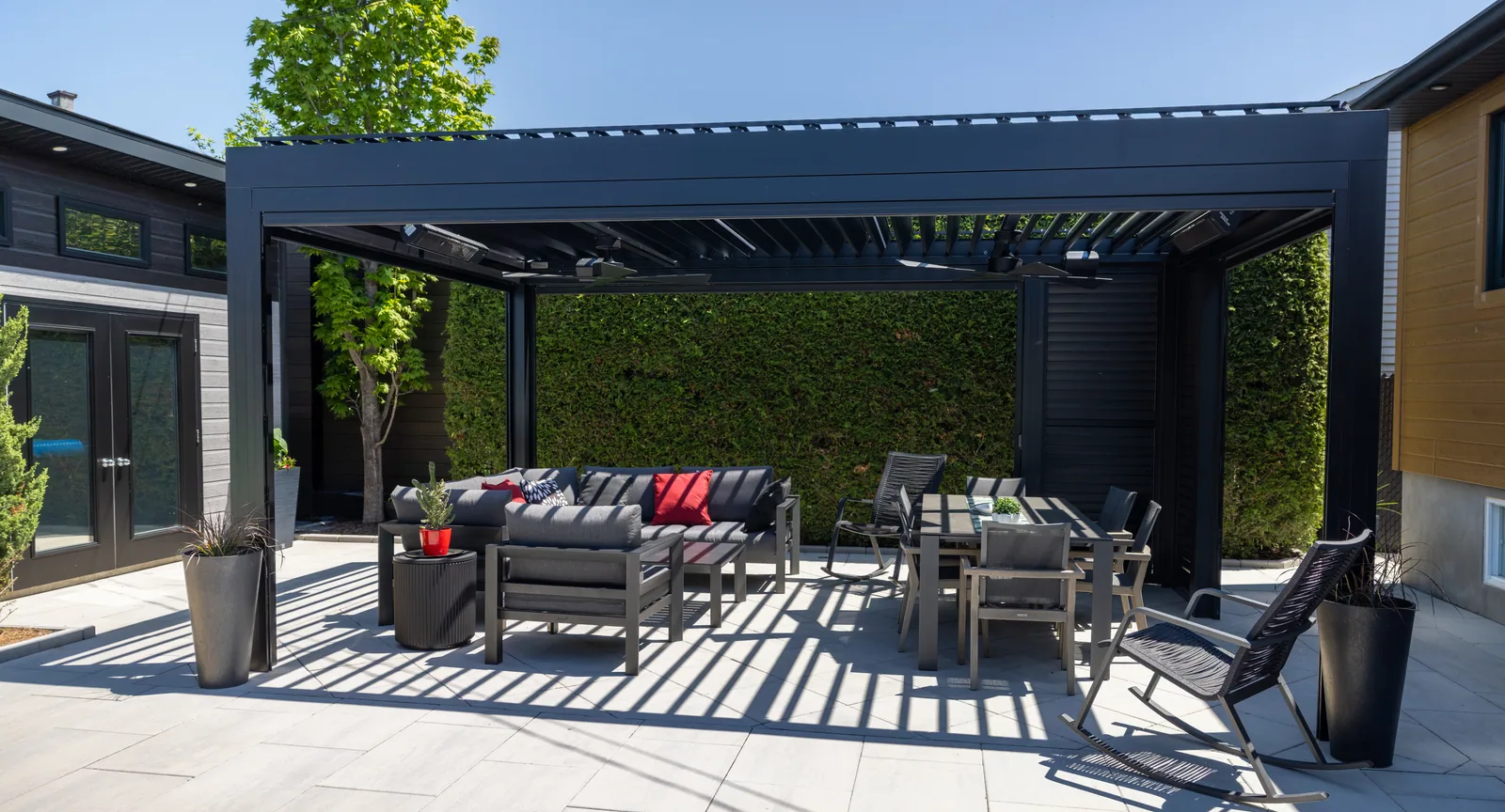 Black Capri bioclimatic pergola over a paved terrace, between the house and a cedar hedge, in Laval