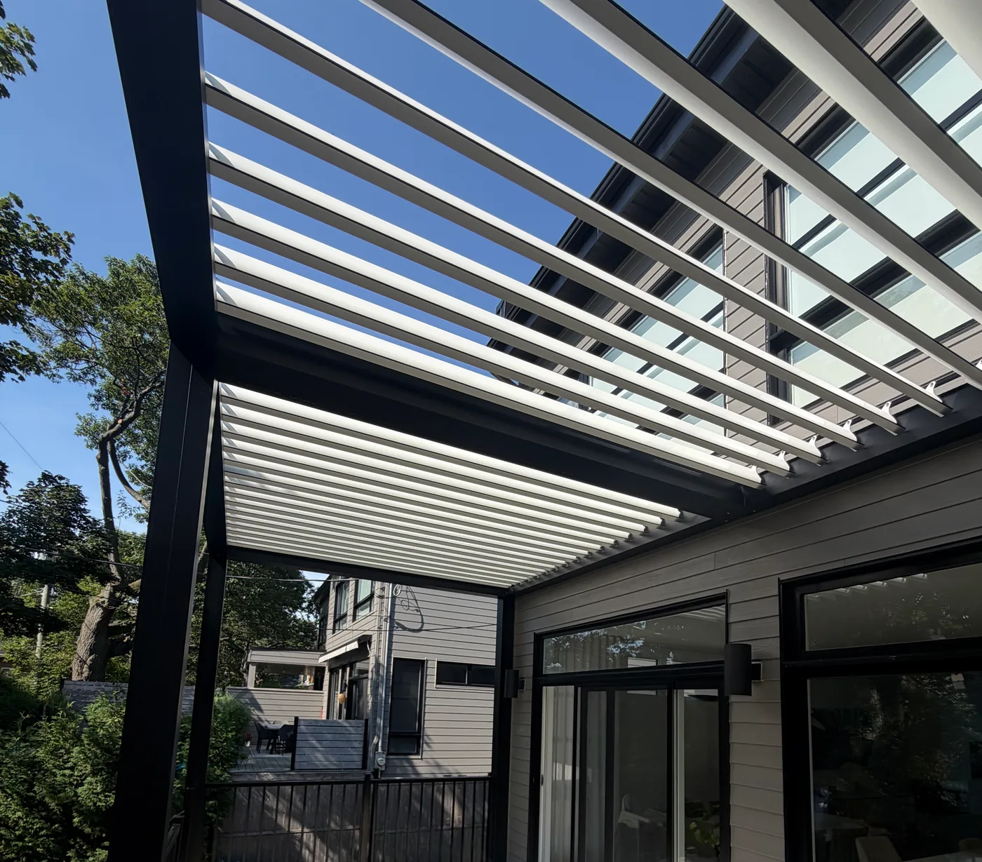 Sous la pergola, lames ouvertes : le ciel paraît entre les lames et le toit rejoint le mur de la maison, au-dessus des portes coulissantes