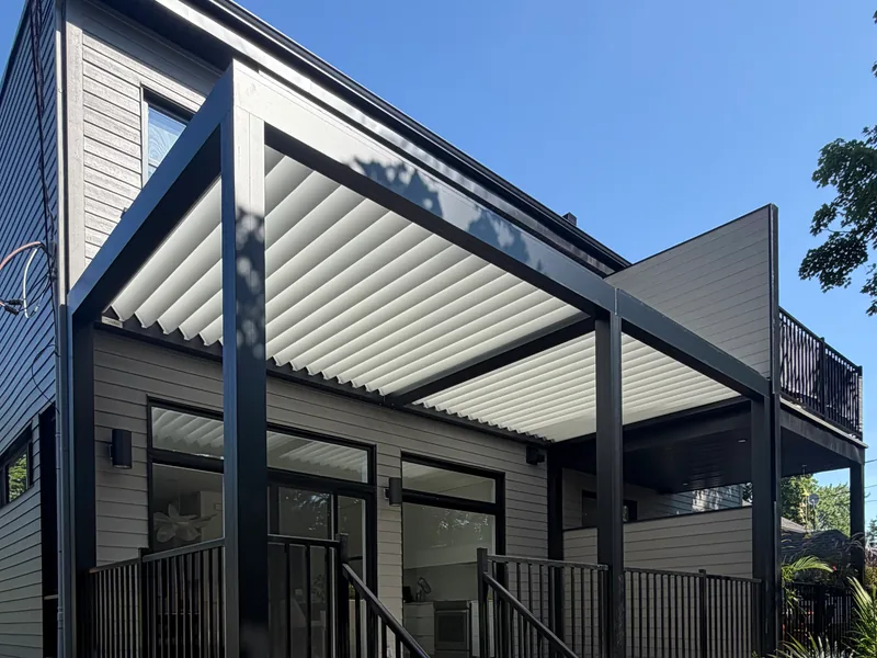 Pergola Capri à lames blanches et structure noire, montée contre une maison contemporaine, au-dessus d’une terrasse surélevée à garde-corps noirs