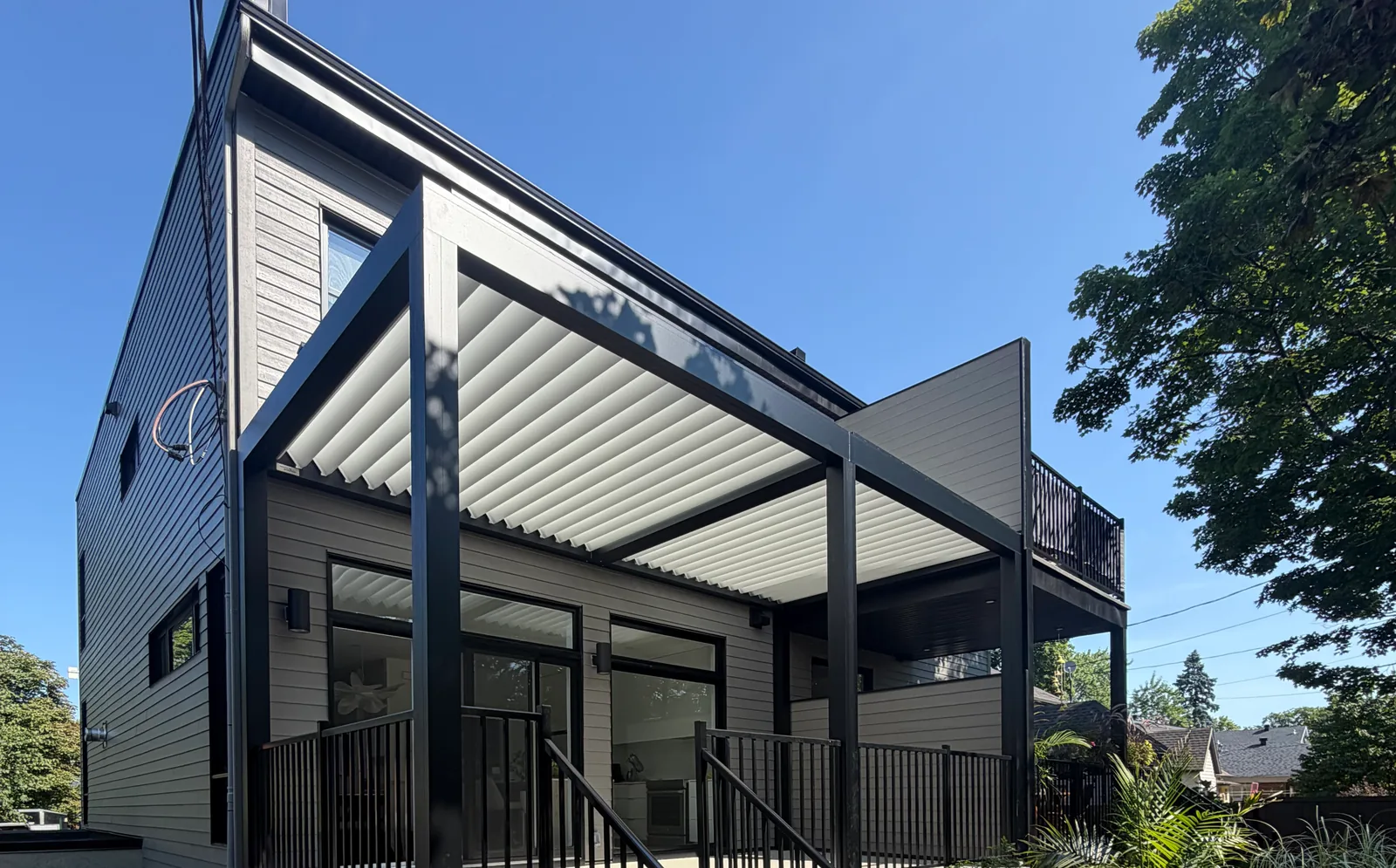 Pergola Capri à lames blanches et structure noire, montée contre une maison contemporaine, au-dessus d’une terrasse surélevée à garde-corps noirs