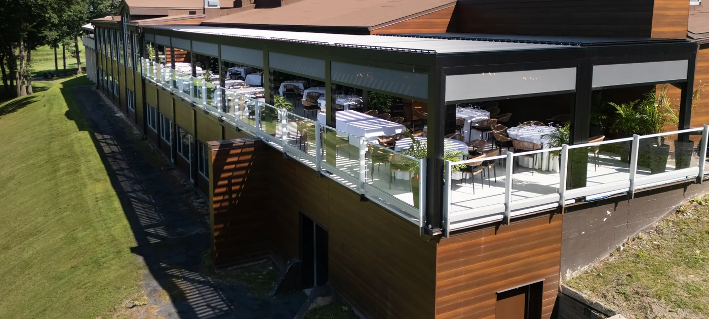 La terrasse vue depuis le bout du bâtiment : les tables sont dressées sous la pergola, le parcours descend à gauche