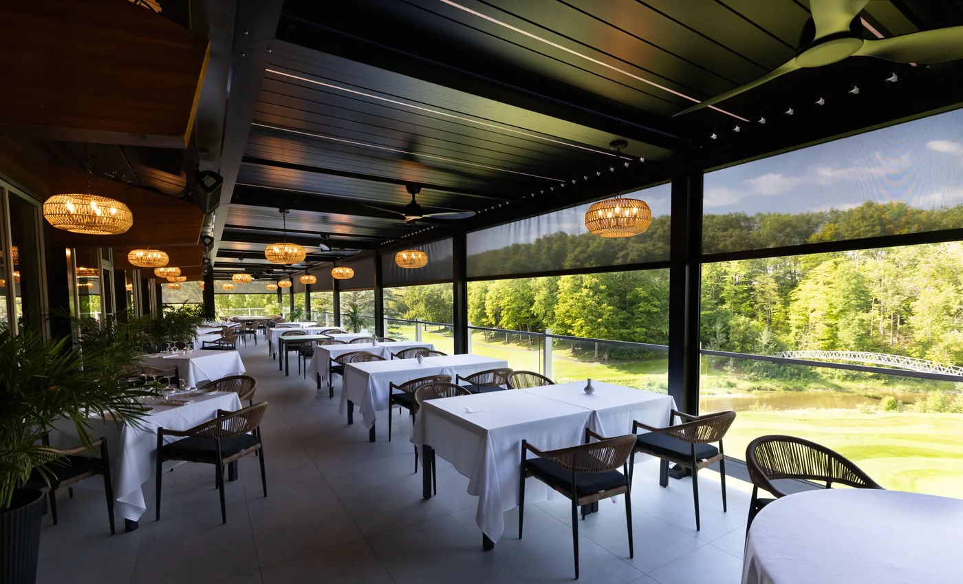 Tables dressées sous la pergola, toiles baissées du côté du parcours, luminaires en rotin fixés aux poutres noires
